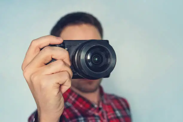 Close up photo of man in hat on blue background taking a photo with digital mirrorless camera. Young cute male photo reporter with a small camera in her hands. Paparazzi taking a picture.