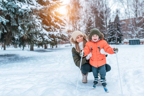 Woman mother helps her child, boy of 4-5 years old, stand children's skis, joy smile fun. First steps skiing. Support, learning healthy lifestyle. In winter, in city park forest. Free space for text. mujer madre ayuda a su hijo, niño de 3-6 años de edad, stand de esquís infantiles, alegría sonrisa diversión. primeros pasos para esquiar. apoyo, aprendizaje de un estilo de vida saludable. en invierno, en el parque forestal de la ciudad. espacio libr