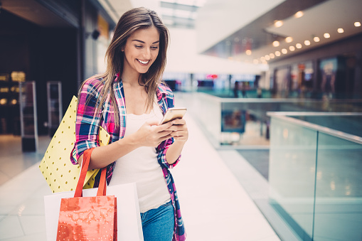 Smiling woman with shopping bags in the clothing store texting on smartphone