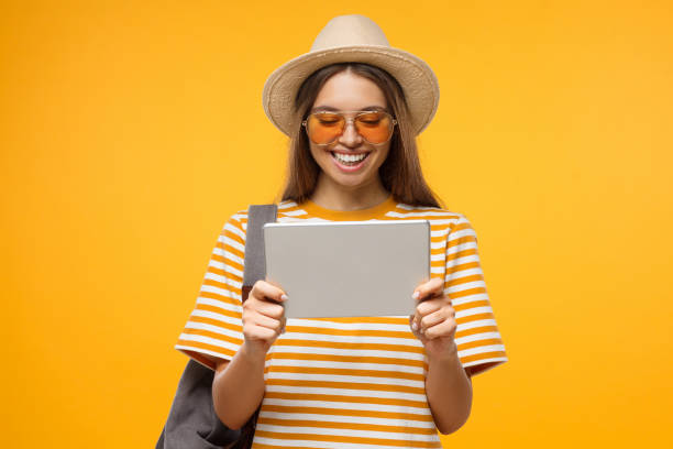 studio portrait of happy smiling young female tourist holding tablet pc, isolated on yellow background - travel ipad isolated backpack imagens e fotografias de stock