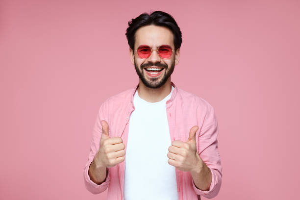 jeune homme attrayant dans la chemise rose, tshirt blanc et lunettes de soleil souriant et montrant le pouce vers le haut le geste sur le fond rose - t shirt shirt pink blank photos et images de collection