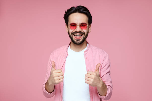 attractive young man in pink shirt, white tshirt and sunglasses smiling and showing thumb up gesture over pink background - t shirt shirt pink blank imagens e fotografias de stock
