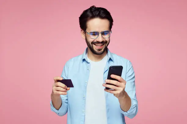 Closeup portrait of young bearded man holding plastic credit card and mobile phone isolated over pink background. Stylish male making shopping on smartphone