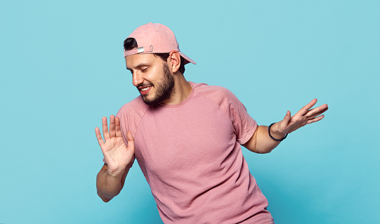 Handsome cheerful boy in stylish clothes and pink baseball cap dancing happily on blue background