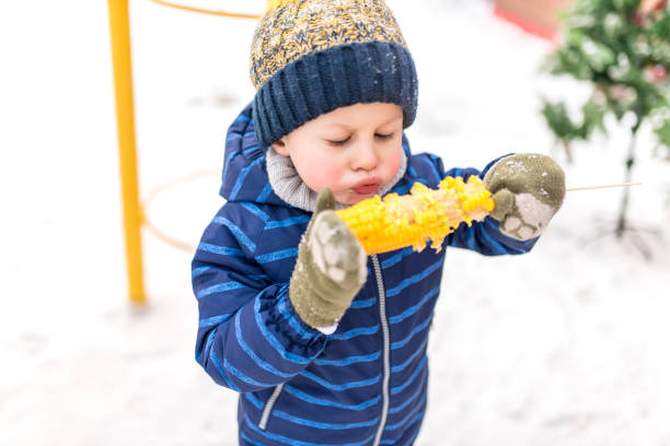 kleine junge 4-5 jahre alt, im winter in der stadt an der frischen luft auf dem hintergrund von schnee. auf heißgekochtem mais gesprengt. frühstück mittagessen und snacks am wochenende an einem kalten festtag. in warmen kleidern und einem hut. - 4 5 years flash stock-fotos und bilder