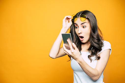 ¡oh mi! Hermosa chica con un teléfono en la mano se sorprende por algo que vio en la pantalla está sosteniendo sus gafas en frente de fondo amarillo brillante. photo