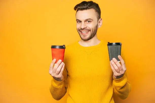 Wonderfully looking man in mustard colored sweater is holding two cups of latte in black and red coffee cups in front of rich yellow background.