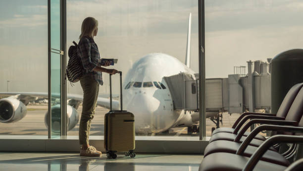 femme avec des cartes d'embarquement et des bagages à main regarde dehors la fenêtre de terminal sur un grand avion de ligne - embarquer photos et images de collection