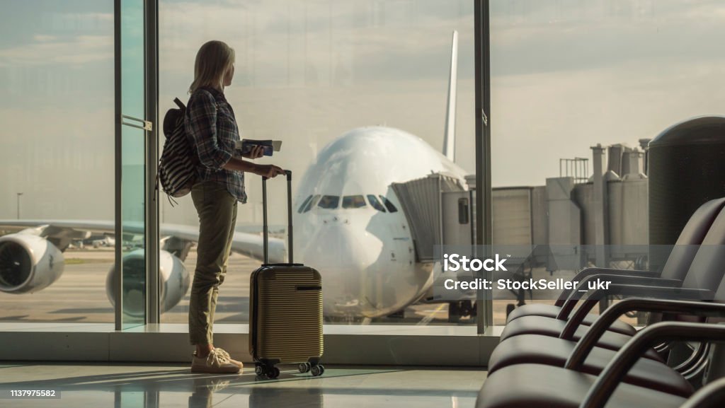 Woman with boarding passes and hand baggage looks out the terminal window on a large airliner Woman with boarding passes and hand baggage looks out the terminal window on a large airlinerStart of the journey. Airport Stock Photo
