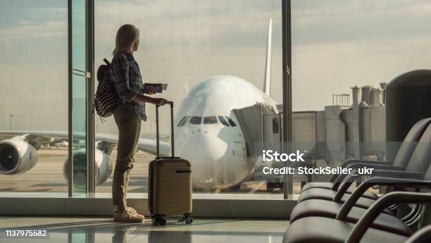 Woman With Boarding Passes And Hand Baggage Looks Out The Terminal Window On A Large Airliner Stock Photo - Download Image Now