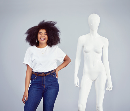 Studio shot of a young woman posing next to a mannequin against a grey background
