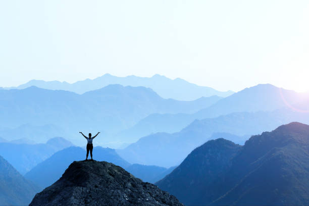 Female Hiker Celebrating Success A female hiker stands on top of a rocky promontory and holds up her arms in celebration in front of a series of mountain ridges that seem to disappear into the distance as the haze obscures the details creating a graphic outline of the various layers of ridge lines. mountain climbing photos stock pictures, royalty-free photos & images