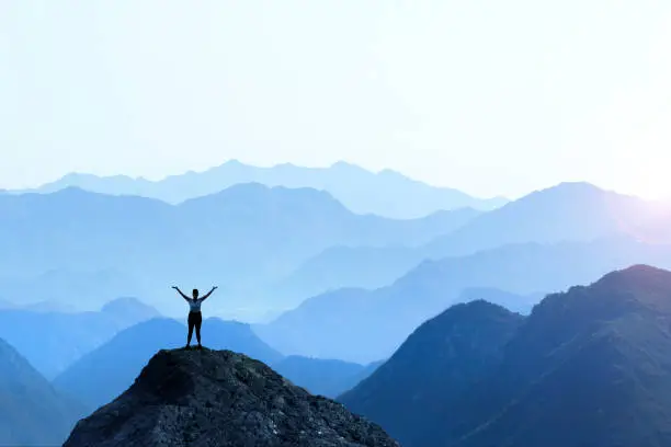 A female hiker stands on top of a rocky promontory and holds up her arms in celebration in front of a series of mountain ridges that seem to disappear into the distance as the haze obscures the details creating a graphic outline of the various layers of ridge lines.