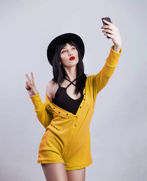 Fashion hipster woman taking selfie and giving peace, victory or two sign. Black hat, yellow knitting overalls, brunette, bright red lips. Trendy fashion style on the gray background Fashion hipster woman taking selfie and giving peace, victory or two sign. Black hat, yellow knitting overalls, brunette, bright red lips. Trendy fashion style on the gray background beautiful women giving head stock pictures, royalty-free photos & images