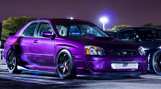Modified Metallic Purple Subaru WRX under streetlights during a local car meet up. Car is parked nose out in parking spot next to other modified cars at the event. New Jersey.
