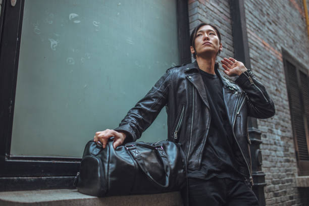 Handsome Asian man with leather jacket and bag standing outdoors.