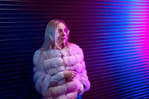 Teen hipster girl in stylish glasses and fur standing on purple street neon light wall background, female teenager fashion model pretty young woman looking at night club city light glow, back to 80s