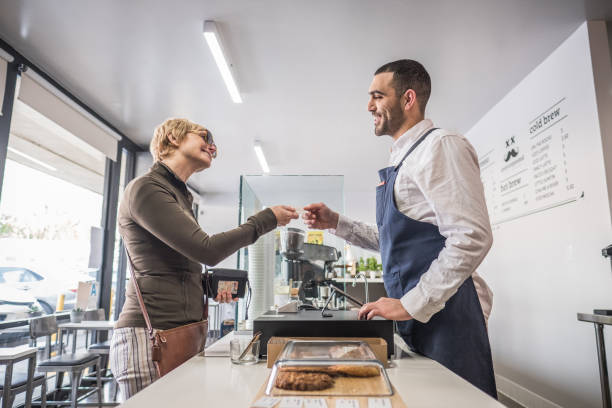 Female Customer Buying Coffee with Credit Card at a Coffee Shop Profile view of a blonde woman handing her  credit card to the employee at the register at a local neighborhood coffee shop. point of sale tablet stock pictures, royalty-free photos & images