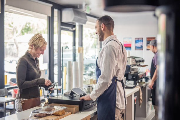 Female Customer Buying Coffee with Credit Card at a Coffee Shop Profile view of a blonde woman reaches into her wallet to give her credit card to the employee at the register at a local neighborhood coffee shop. point of sale tablet stock pictures, royalty-free photos & images