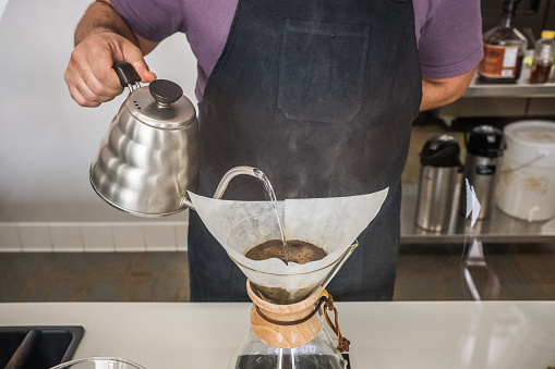 Close up of barista pouring boiling water from a metal teapot into a glass carafe to make a pour over coffee at a coffee shop.