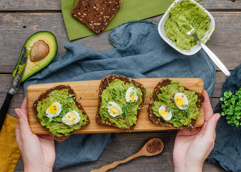 Female hands holding homemade sandwiches with avokado, quail eggs and radish sprouts on rustic wooden background. Top view, flat lay.
