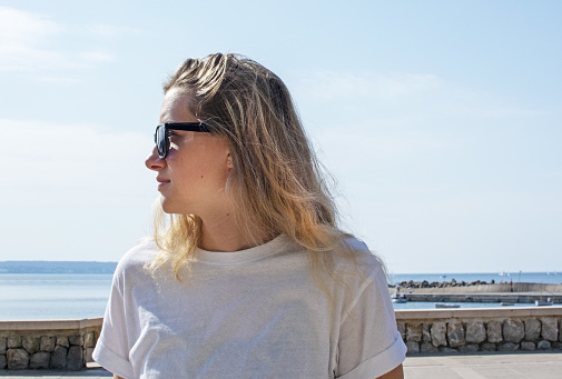 Young casual sporty dressed blonde woman with sunglasses in white t-shirt sits near street with ocean behind in Mallorca, Spain.