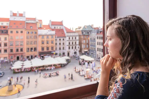 Young woman sitting looking outside through window sill of apartment with view of old market square in town of Warsaw, Poland dreaming