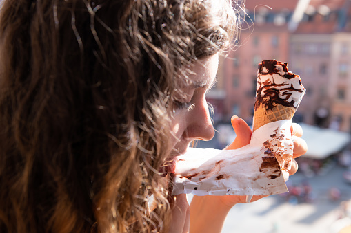 Woman eating chocolate ice cream gelato cone paper licking with background of Warsaw, Poland old market square historic buildings in city during sunny summer day