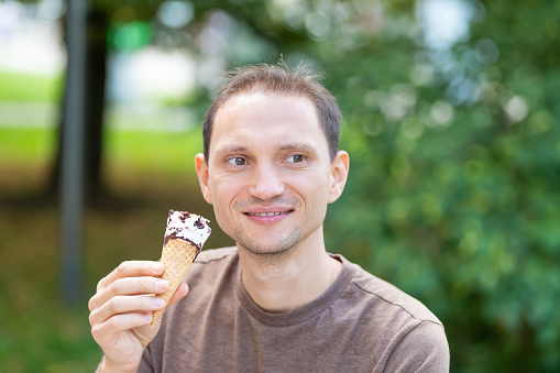Man happy smiling holding one vanilla chocolate ice cream gelato cone with bokeh background of park in Europe city during sunny summer day