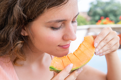 Young woman in white summer smiling eating orange cantaloupe melon slice outside in Italy villa sunny sunlight