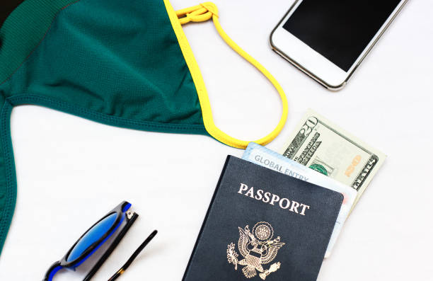 Packing for overseas summer holiday: a woman's essentials laid out on a bed (swimsuit, smart phone, US passport, global entry card, us dollars, sunglasses).