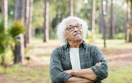 A senior Hispanic man in his 80s standing outdoors in a park, trees out of focus in the background. He has white hair and is wearing eyeglasses and a plaid shirt.