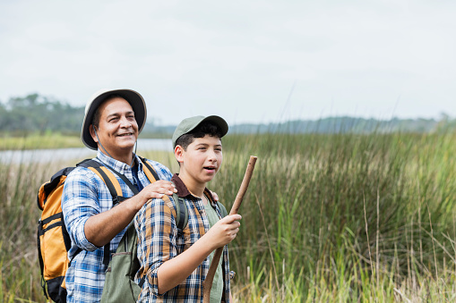 An Hispanic 12 year old boy and his father, a mature man in his 50s, hiking in a park, exploring nature. They are standing and looking out toward wetlands, a grassy area by water, enjoying the view.