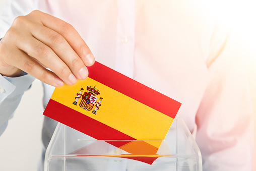 Human hand is inserting Spain flag into ballot box.