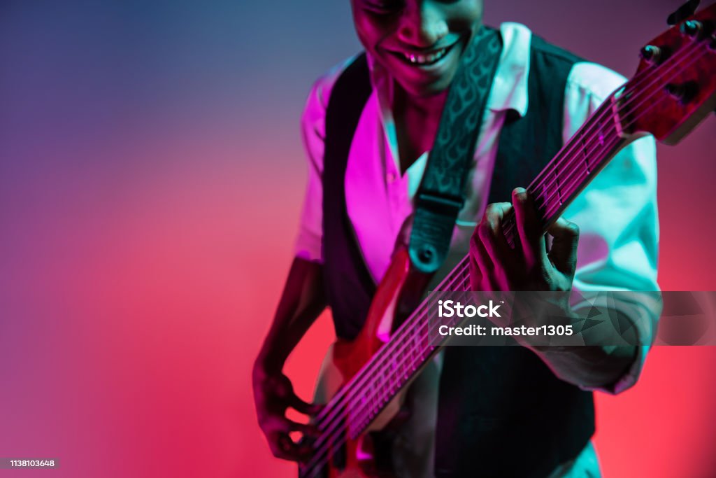 African American jazz musician playing bass guitar. African American handsome jazz musician playing bass guitar in the studio on a neon background. Music concept. Young joyful attractive guy improvising. Close-up retro portrait. Musician Stock Photo