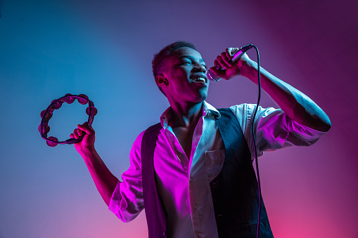 African American handsome jazz musician playing tambourine and singing into the microphone in the studio on a neon background. Music concept. Young joyful attractive guy improvising. Close-up retro portrait.