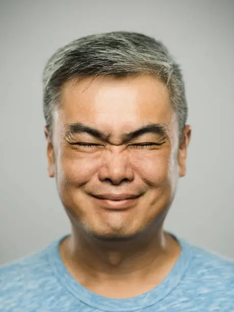 Close up portrait of mature asian man with disgusted expression and eyes closed against gray white background. Vertical shot of chinese real people grimacing in studio with gray hair. Photography from a DSLR camera. Sharp focus on eyes.