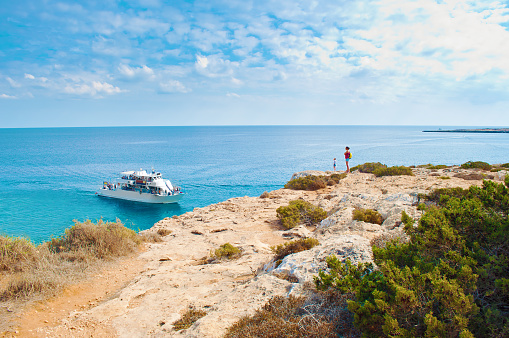 Two tiny figures of girls standing on the edge of a cliff. One white tourist ship in a bay near Cape Greco, Cyprus. Rock coastline near deep green transparent azure water. Warm day in fall