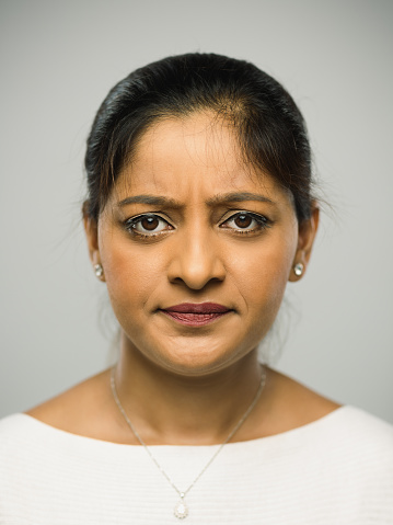 Close up portrait of indian young woman with upset expression against gray background. Vertical shot of indian real people frowning in studio with long black hair. Photography from a DSLR camera. Sharp focus on eyes.