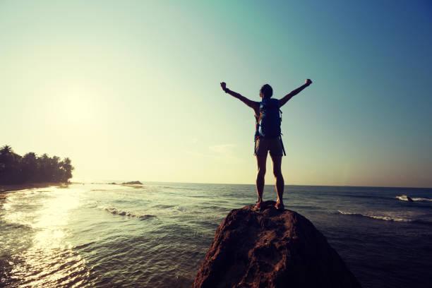 istock Successful young woman with backpack outstretched arms on seaside rock cliff edge 1138127362