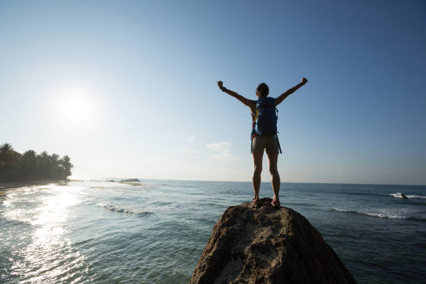 istock Successful young woman with backpack outstretched arms on seaside rock cliff edge 1138127447