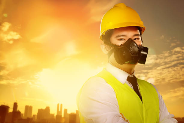 Handsome asian worker with protective mask and yellow helmet standing with sunset on the city background. Labor day concept