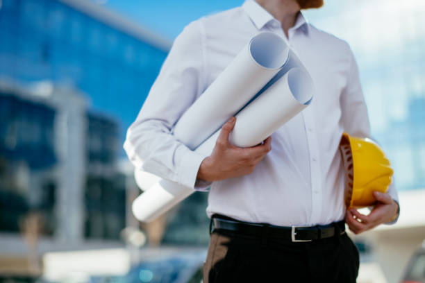 Young male architect holding a yellow helmet and blueprint. Shadow DOF. Developed from RAW; retouched with special care and attention; Small amount of grain added for best final impression. 16 bit Adobe RGB color profile.