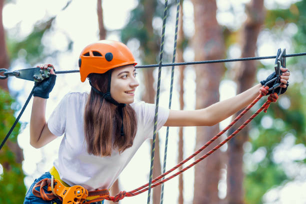 Young beauty female woman adult wears protective helmet having fun in extreme rope park, amusement park. Climbing in rope bridge at green nature forest. Active healthy lifestyle in spring or summer.