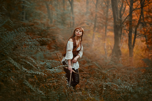 gorgeous lady with long red hair in leather clothes follows wild animal, hunts down prey in rainforest, rite of initiation into hunters, girl pulled bowstring of bow with arrow, ready to shoot.