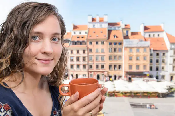 Young woman sitting on window sill windowsill with teacup holding tea coffee mug cup in old market square in town of Warsaw, Poland