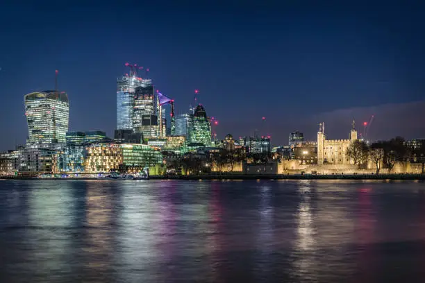 City of London's financial district and Tower of London famous landmark and tourist attraction during night illumination as photographed from Southwark bank. Long exposure technique used to blur the river Thames high tide waters reflecting the city lights. Shot on EOS R full frame system with premium RF lens for highest quality.