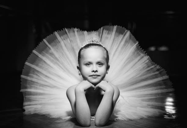 foto en blanco y negro de una linda bailarina sonriente en tutu blanco y una corona que se pone en el suelo con las manos debajo de la barbilla. - ballet dress studio shot costume fotografías e imágenes de stock