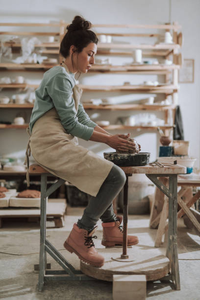 Beautiful young lady in apron working on pottery wheel Vertical full length portrait of charming craftswoman kneading and shaping clay in pottery workshop timberland arizona stock pictures, royalty-free photos & images