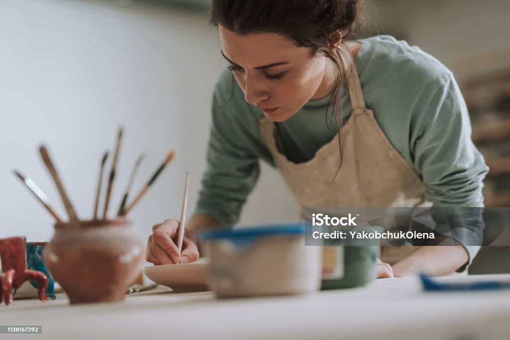 Skillful young woman in apron painting pottery at workshop Close up portrait of beautiful brunette lady drawing on clay plate while leaning over the table with pottery and paints Painting - Activity Stock Photo